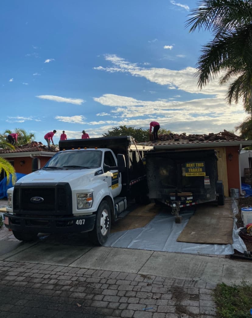 Roofing crew working on house with truck and trailer parked nearby under cloudy sky.