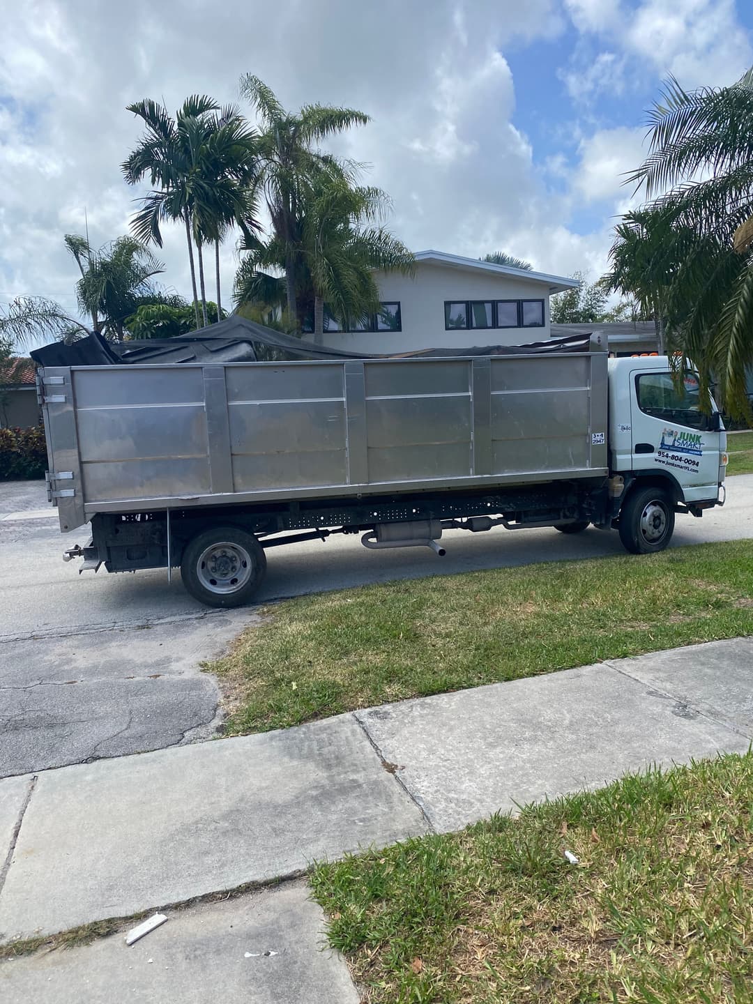 Silver dump truck parked on a residential street with palm trees and a house in the background.