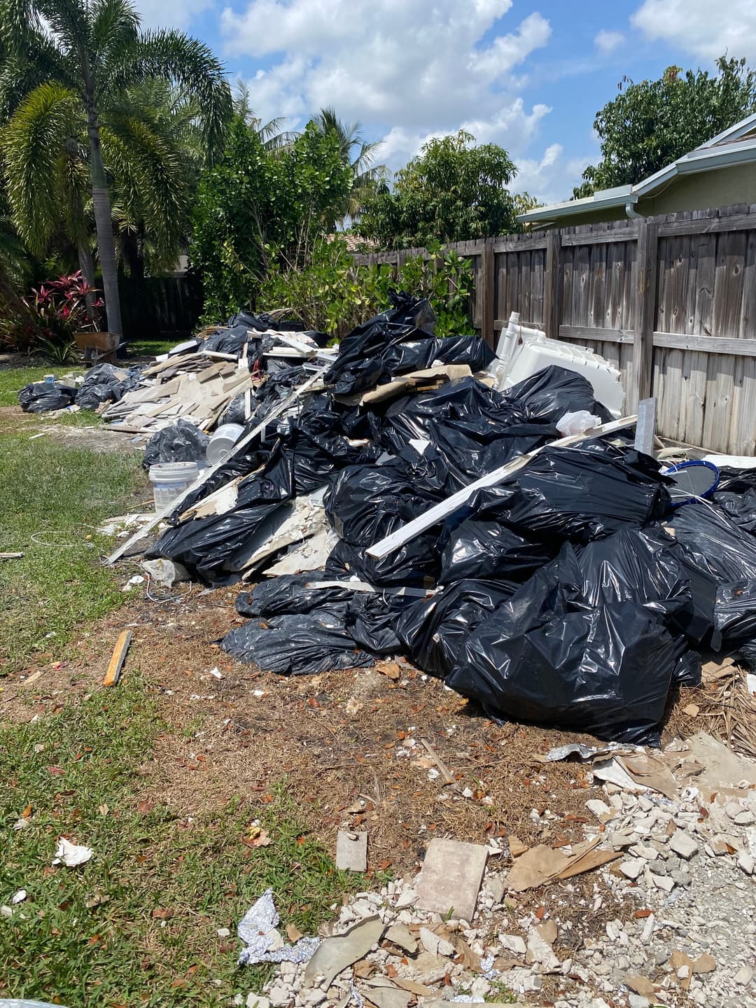 Piled garbage bags and debris in a neglected yard with palm trees and wooden fence.