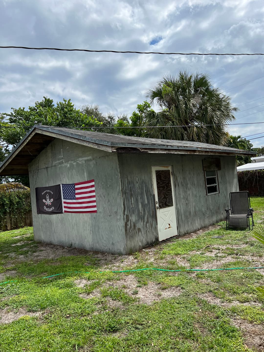 Weathered shed featuring a U.S. flag and pirate flag, surrounded by greenery and palm trees.
