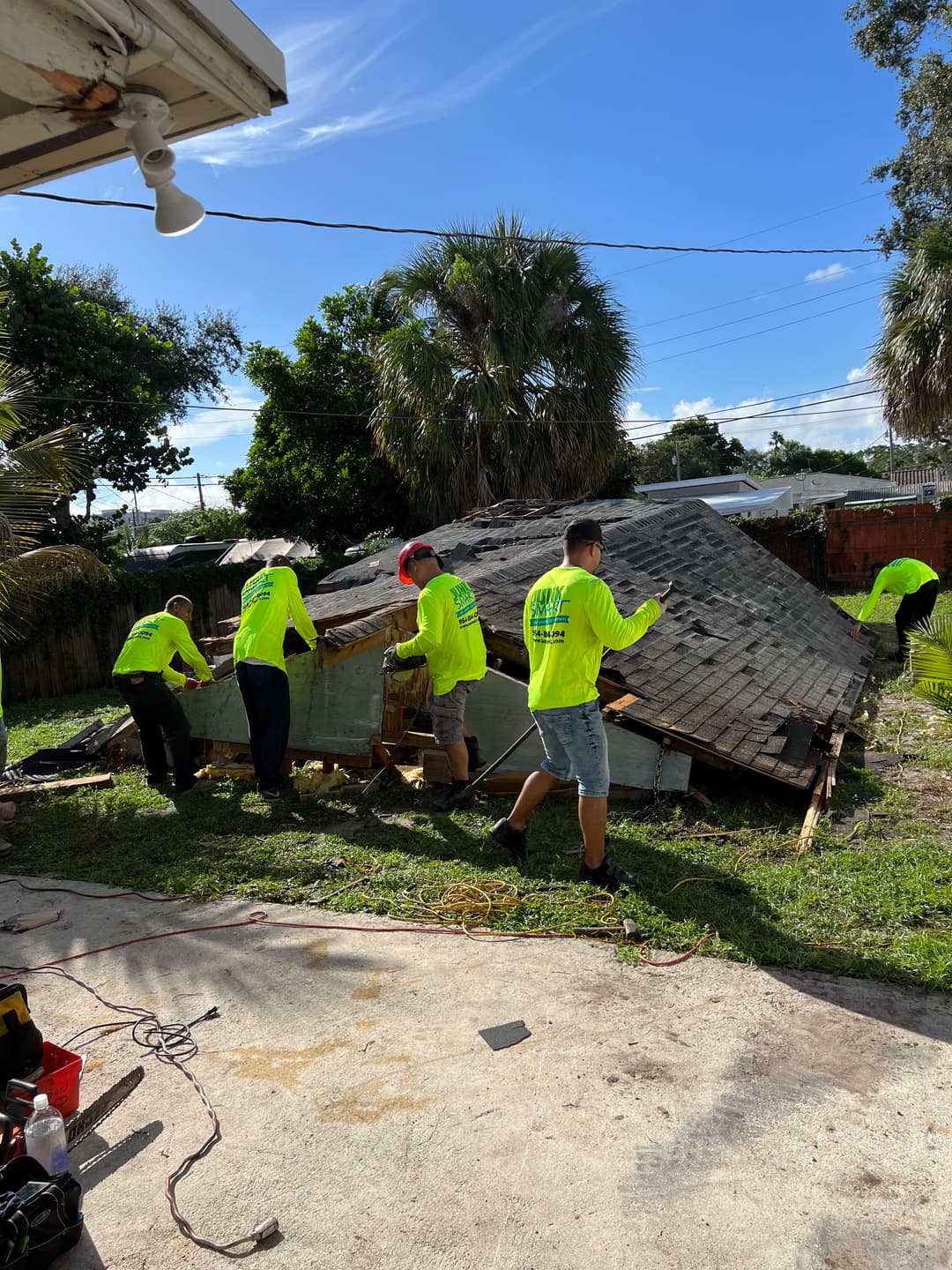 Team of workers in bright shirts repairing a collapsed roof in a tropical backyard.