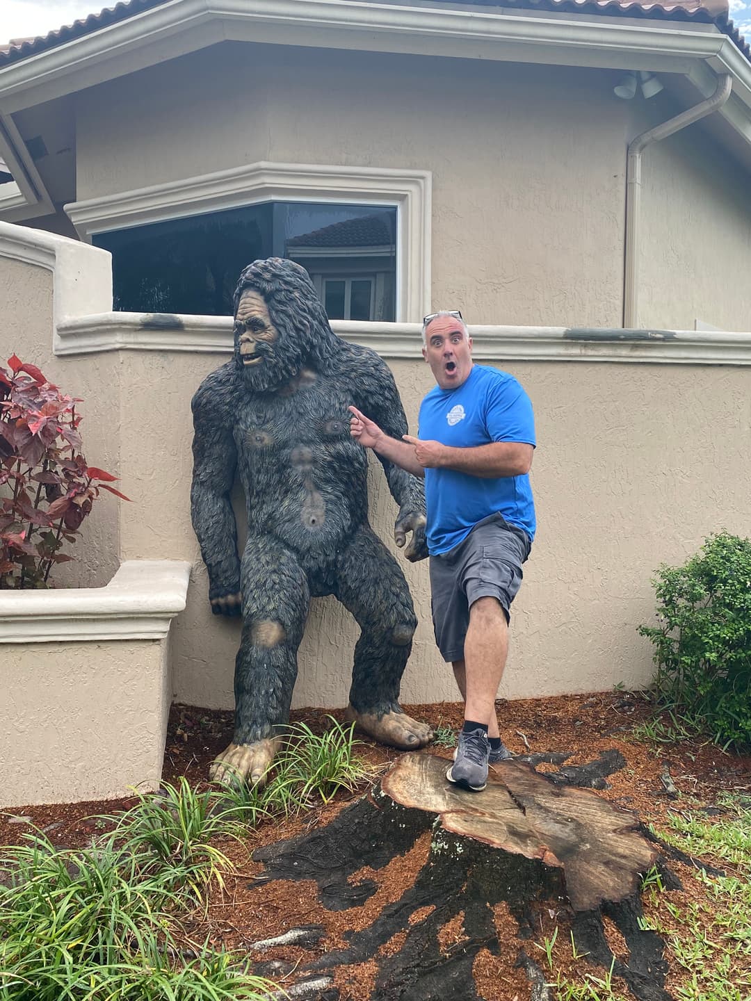 Person posing excitedly next to a life-sized Bigfoot statue outside a house.