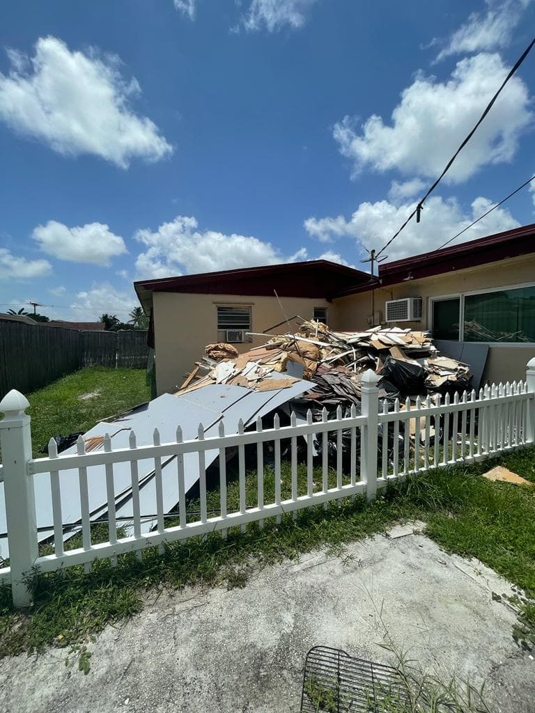 Debris pile outside a house with a white fence under a blue sky. Cleanup needed.