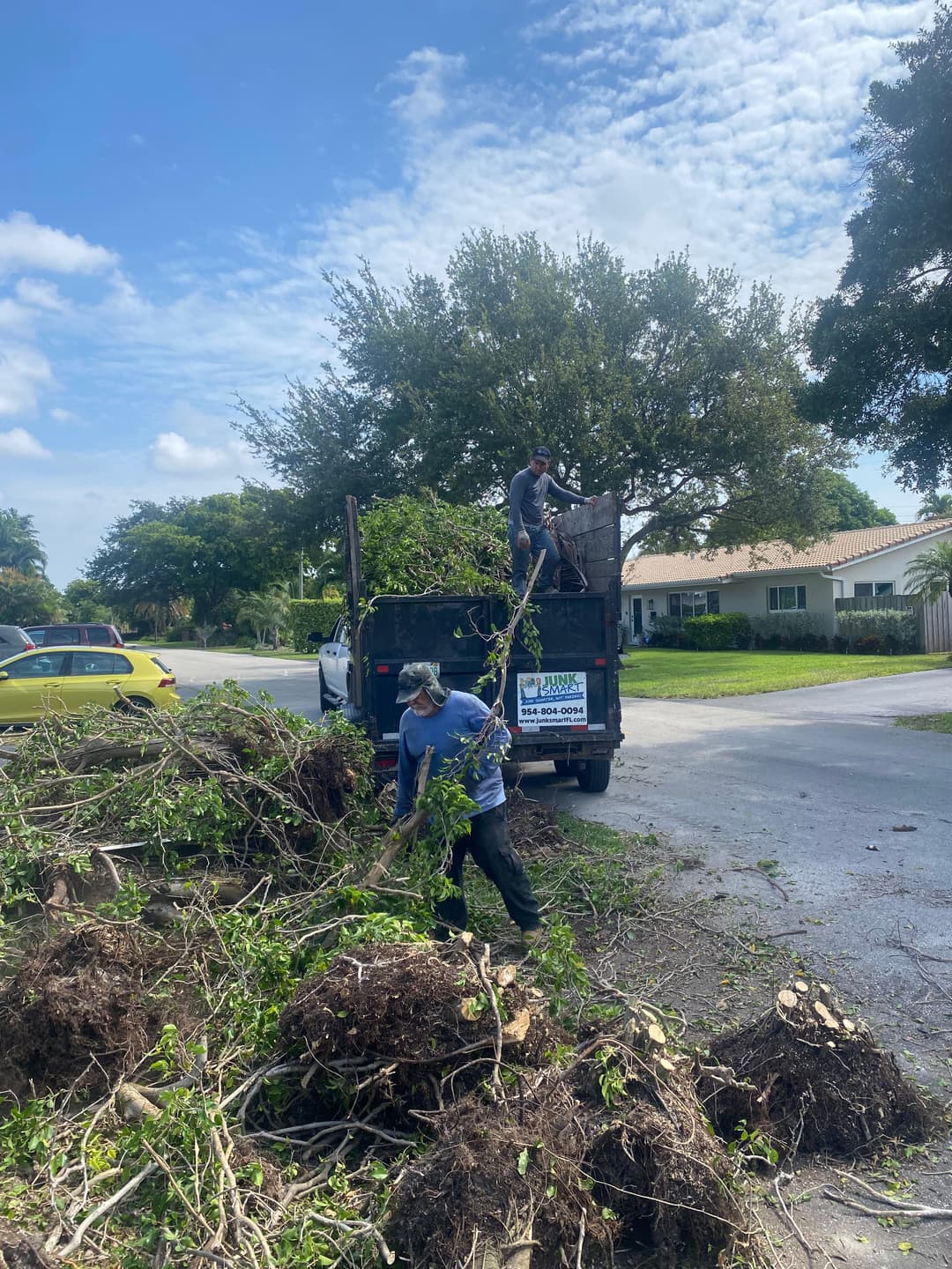 Workers clearing yard debris into a truck on a sunny day, with trees and a home in the background.
