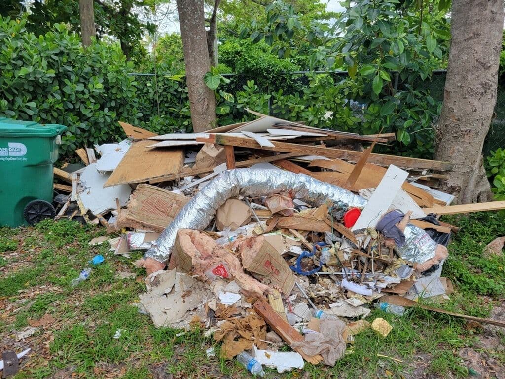 Pile of construction debris and trash near green dumpster amidst lush vegetation.