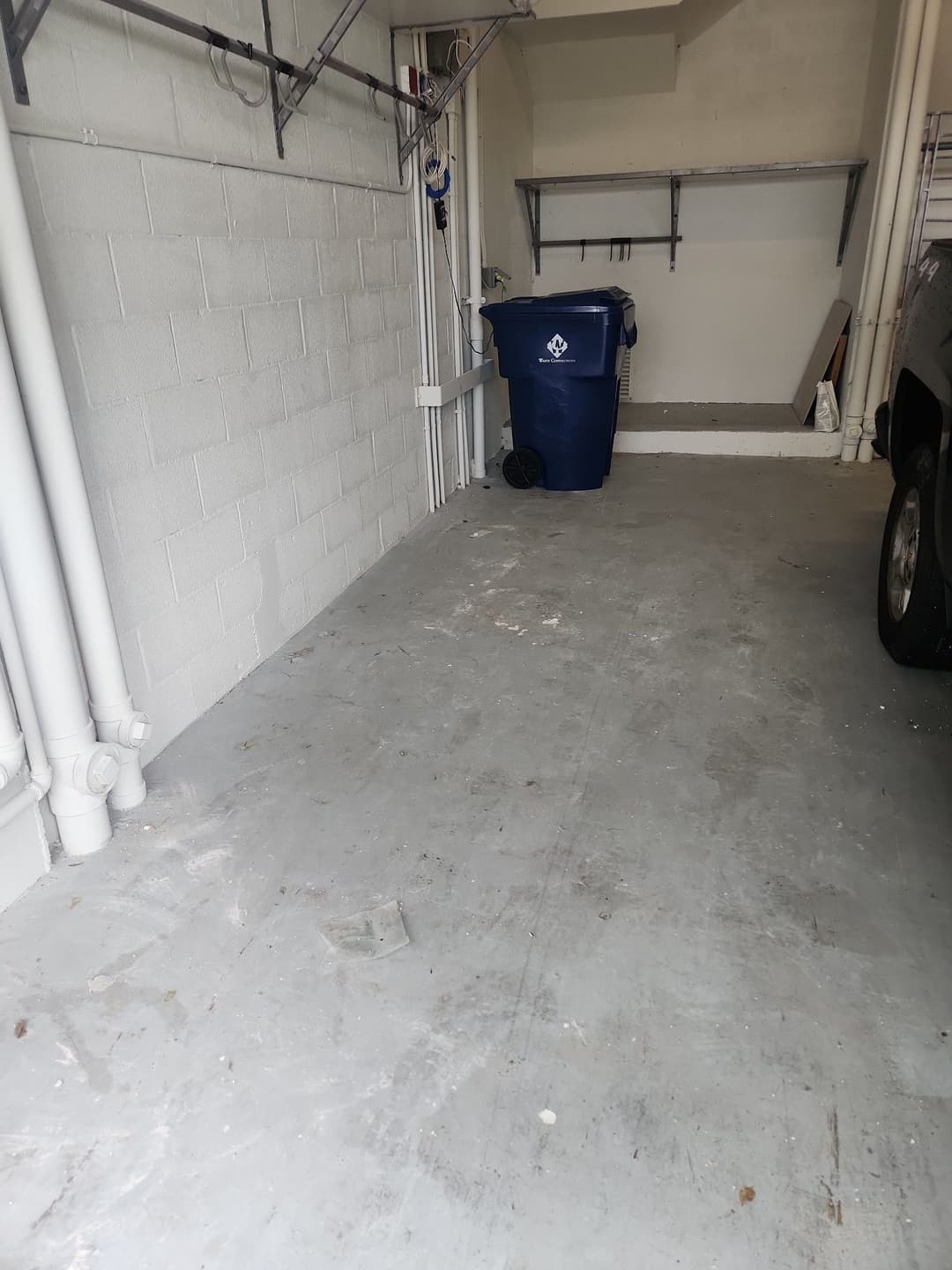 Empty garage space with gray flooring, white walls, and a blue trash bin.
