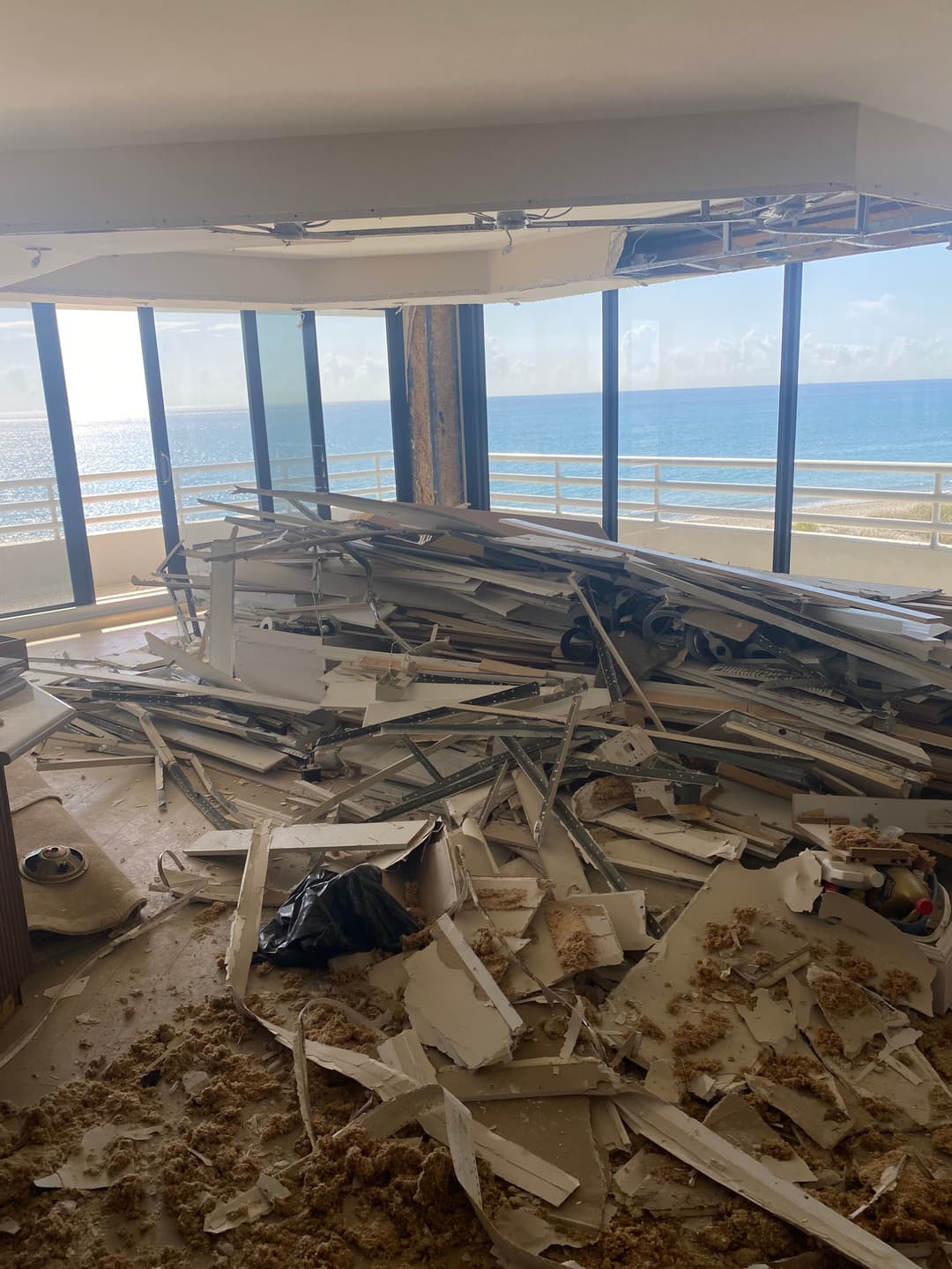 Interior of a damaged room with debris and ocean view through large windows.