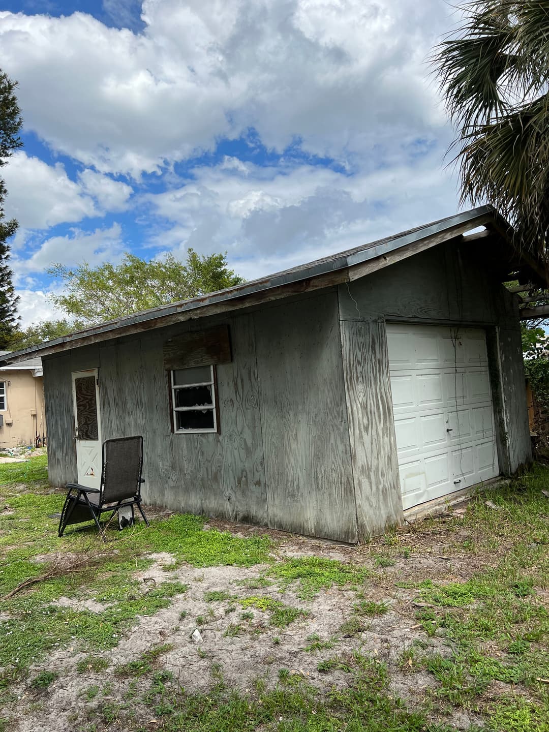 weathered shed with garage door in a grassy yard under a cloudy sky. Chair in foreground.
