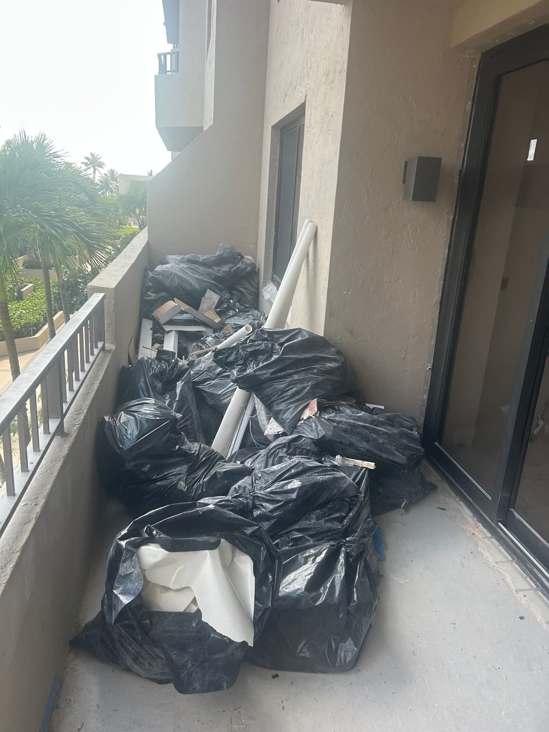 Black garbage bags piled on a balcony, with outdoor view and tropical plants visible.