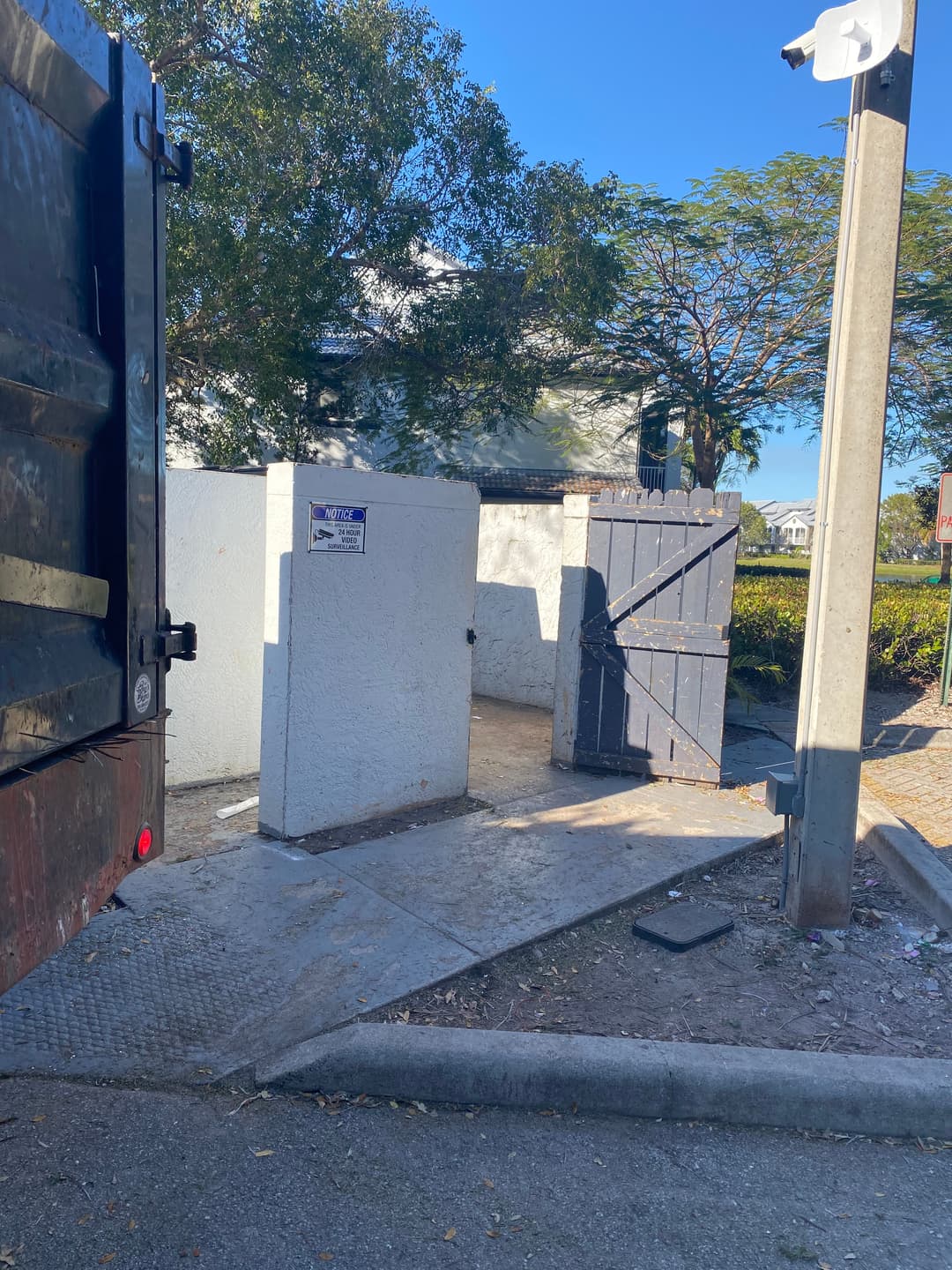 Trash bins near a street with a wooden gate and surrounding trees on a sunny day.