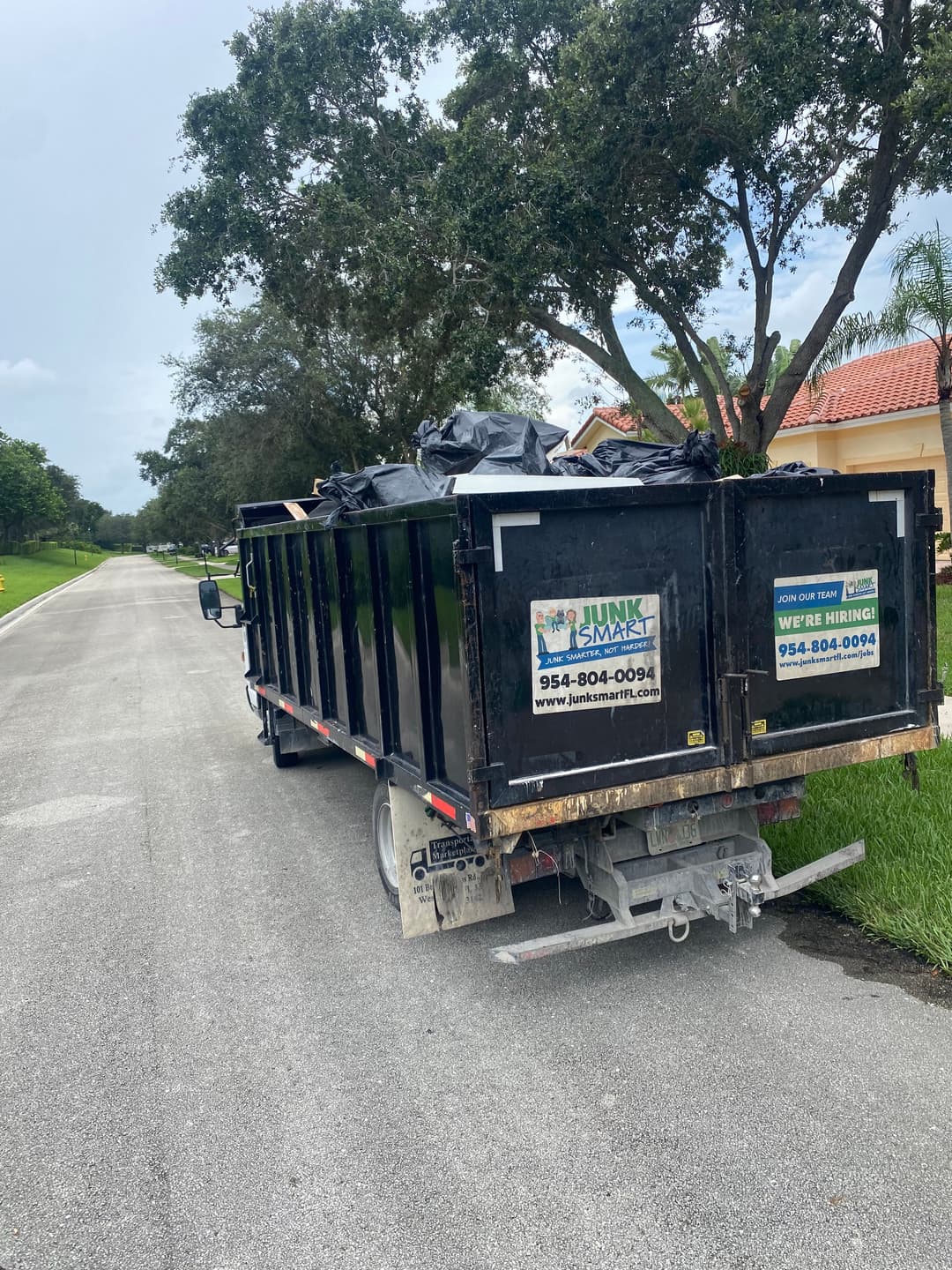 Black dumpster on a residential street, filled with debris, promoting Junk Smart services.